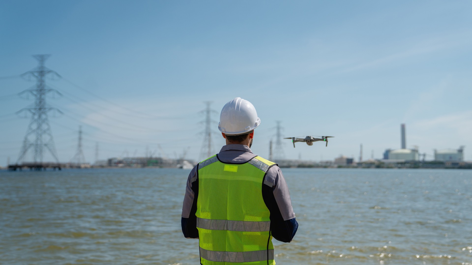 Engineer in a safety vest operating a drone to inspect and survey electrical power lines. Technician inspecting the power station for scheduled maintenance.