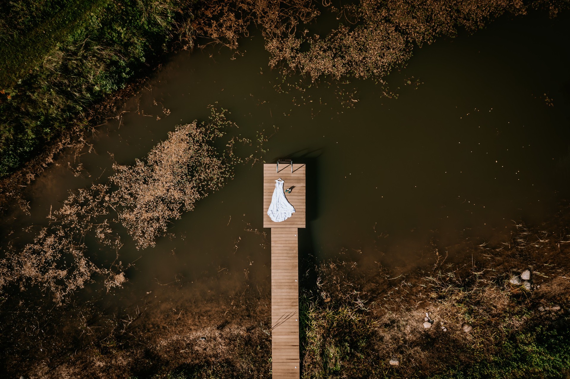 A white wedding dress and shoes are placed on a wooden dock over a murky pond surrounded by greenery, captured from above with ample copy space.