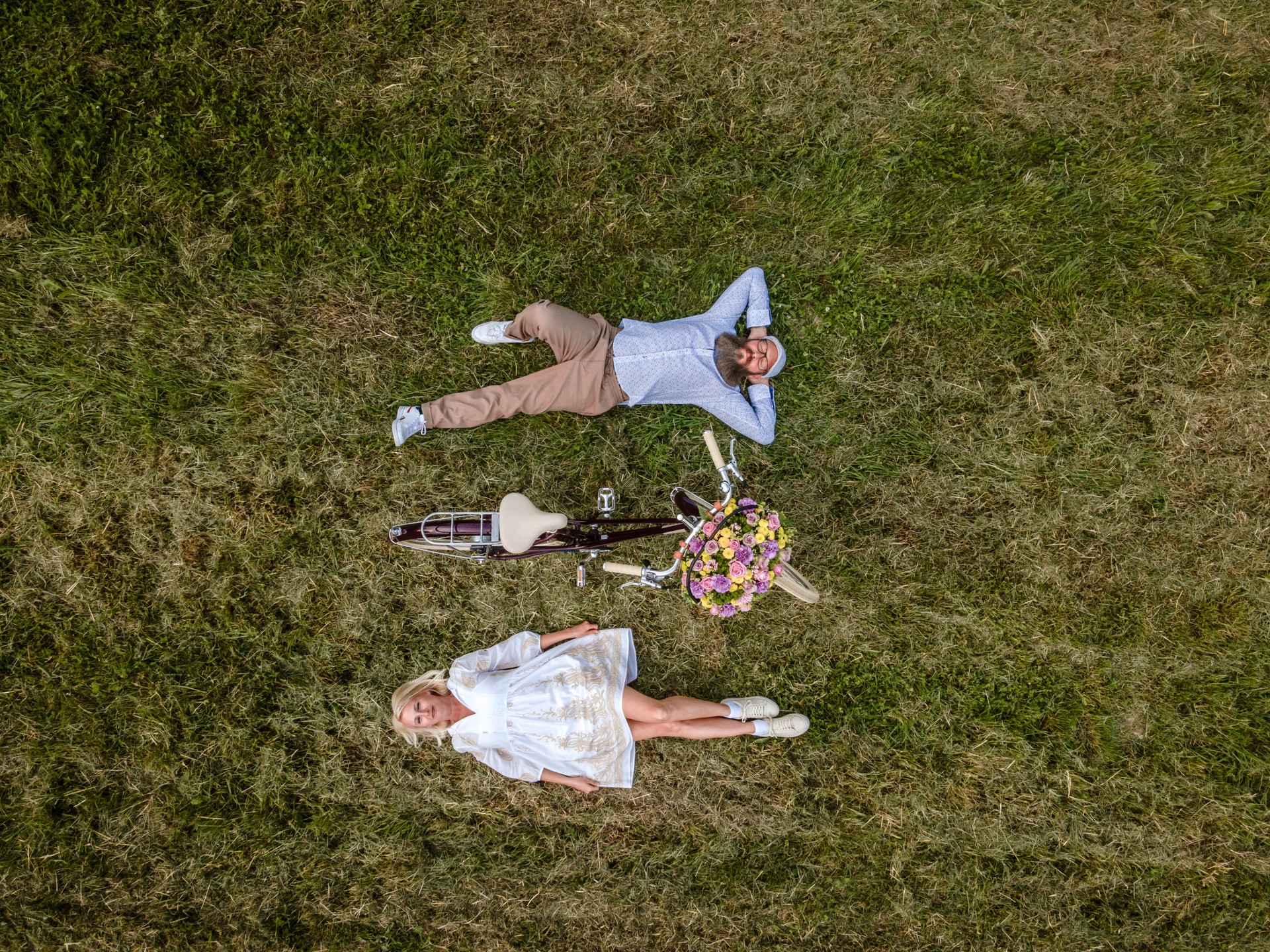 aerial drone shot of blonde woman with white dress and her boyfriend or husband posing with bicycle with beautifully decorated flower basket in nature, nature concept, flowers, delivery service